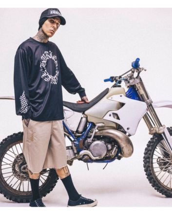 Travis Barker con camiseta de manga larga negra Check Out Jersey y gorra, posando junto a una moto de cross