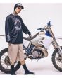 Travis Barker con camiseta de manga larga negra Check Out Jersey y gorra, posando junto a una moto de cross
