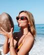 Mujer con gafas de sol I-SEA Carly Havana Tortoise-Marrón Polarized sosteniendo un coco frente al mar